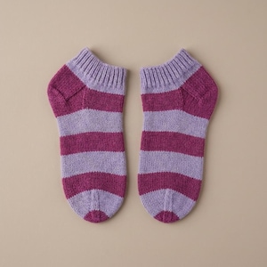 Könnte beinhalten: Ein Paar Knöchelsocken mit horizontalen Streifen in abwechselnden Farbtönen von Magenta und hellem Lavendel. Die Socken verfügen über ein geripptes Bündchen am oberen Rand und einen verstärkten Fersenbereich mit einer gleichmäßigen Strickstruktur.