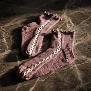May include: A pair of mauve-colored knitted socks with a white braided design down the side. The socks have a ruffled edge at the top and are displayed on a marble surface.