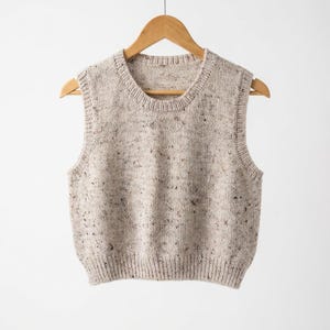 Op de afbeelding: Een beige mouwloze gebreide vest met een gespikkeld patroon, hangend aan een houten hanger. Het vest heeft een geribbelde zoom en halslijn, en de stof lijkt een zachte brei te zijn. De stijl is casual en comfortabel.