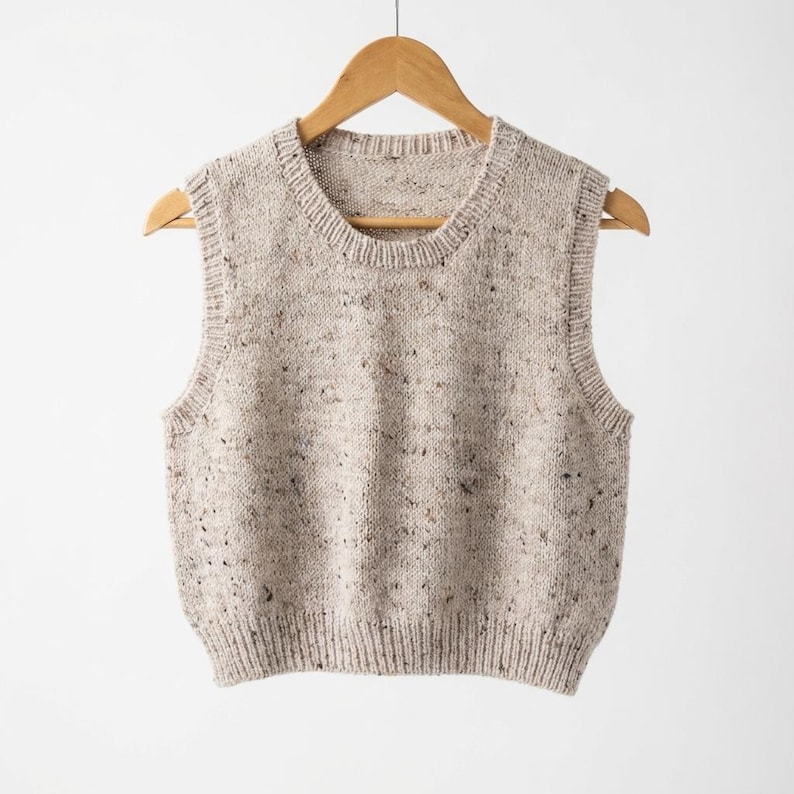 Op de afbeelding: Een beige mouwloze gebreide vest met een gespikkeld patroon, hangend aan een houten hanger. Het vest heeft een geribbelde zoom en halslijn, en de stof lijkt een zachte brei te zijn. De stijl is casual en comfortabel.