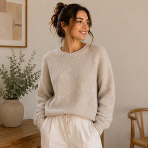 Puede incluir: Un suéter de punto beige de manga larga con cuello redondo y puños acanalados, combinado con pantalones de color blanco roto con cintura de cordón. El suéter tiene un corte relajado y una textura suave y uniforme en toda la prenda.
