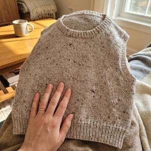 Op de afbeelding: Een beige gebreide vest met een ronde hals en geribbelde zoom. Het vest heeft een gespikkeld patroon en is gemaakt van zacht, getextureerd garen. De mouwloze top is neutraal van kleur.