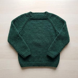 May include: A dark forest green long-sleeve sweater featuring a classic crew neckline, raglan sleeves, and ribbed knit detailing along the collar, cuffs, and hem. The garment is constructed with a chunky knit texture throughout the body and sleeves.