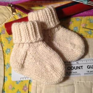 May include: A pair of cream-colored hand-knitted baby booties with a ribbed cuff design. The booties are positioned next to a white ruler showing measurements in inches, indicating the small scale of the knitted footwear items.