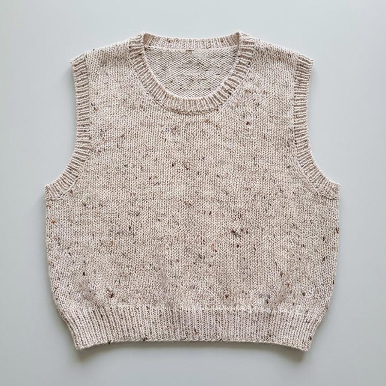 Op de afbeelding: Een beige gebreide vest met een gespikkeld patroon. Het mouwloze kledingstuk heeft een geribbelde zoom, halslijn en armsgaten. De neutrale kleur en de getextureerde brei suggereren een gezellige, handgemaakte esthetiek, geschikt om in laagjes te dragen.