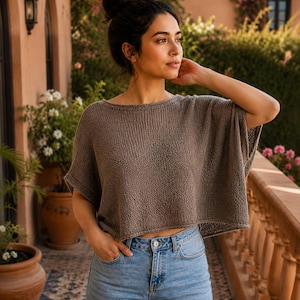 May include: A taupe-coloured, short-sleeved, cropped knit sweater with a wide boat neckline and a loose, boxy fit, paired with light blue denim jeans featuring a button fly and a classic five-pocket design with visible metal rivets.