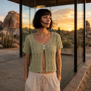May include: A light green short-sleeved knit cardigan featuring a textured openwork pattern and two small white buttons at the neckline. The garment has a scoop neck and a cropped length, paired with light beige drawstring trousers.