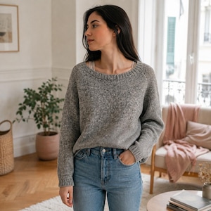Puede incluir: Un suéter de punto gris jaspeado de manga larga con un cuello redondo ancho y puños acanalados. El suéter presenta una textura moteada en toda la tela y se combina con jeans de mezclilla azul claro, creando un estilo casual y neutro.