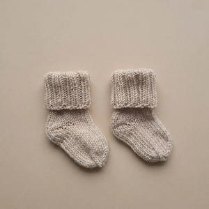 May include: A pair of beige knit socks featuring a thick, ribbed cuff at the top. The socks are crafted with a visible stockinette stitch pattern throughout the foot and heel area, showcasing a neutral, light tan color and a soft, textured appearance.