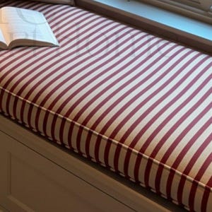 May include: A window seat with a red and white striped cushion and three pillows. The pillows are white, tan, and cream-colored. An open book rests on the cushion. The window looks out onto a winter scene.