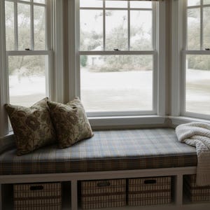 K&ouml;nnte beinhalten: Eine Fensterbank mit einem karierten Kissen und zwei floralen Kissen. Der Sitz hat einen eingebauten Stauraum mit drei gestreiften K&ouml;rben. Eine wei&szlig;e Decke ist auf dem Sitz drapiert. Die Fenster bieten einen Blick auf eine grasbewachsene Fl&auml;che.