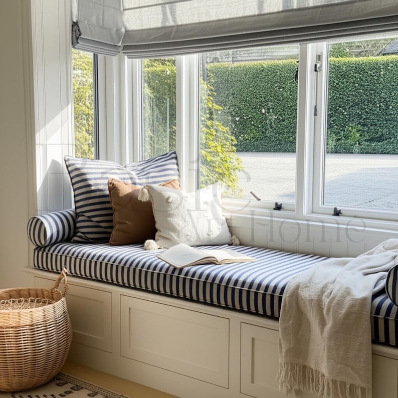 May include: A window seat with blue and white striped cushions and pillows. A brown pillow and a white pillow with an anchor design are also present. A woven basket and a blanket are visible. An open book rests on the seat.