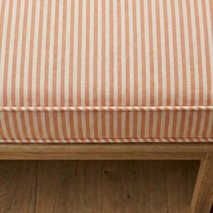 Op de afbeelding: Close-up van een gestreept kussen in perzik en wit. Het kussen maakt deel uit van een houten frame, met verticale strepen. Het kussen heeft een op maat gemaakte rand en het houten frame is zichtbaar aan de onderkant van de afbeelding.