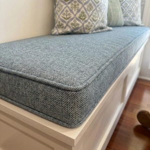 May include: A light blue bench cushion with a textured fabric and a contrasting border. The cushion sits on a white bench with a paneled design. Decorative pillows with floral patterns rest on the bench.