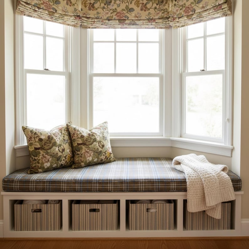 K&ouml;nnte beinhalten: Eine gem&uuml;tliche Fensterbank mit kariertem Kissen und floralen Kissen. Der Sitz hat einen eingebauten Stauraum mit gestreiften Boxen. Eine cremefarbene Strickdecke liegt auf dem Sitz. Ein floral gemusterter Fensterbehang h&auml;ngt &uuml;ber den Fenstern.