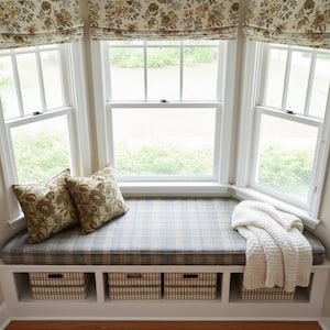 May include: A cozy window seat with a plaid cushion, two floral pillows, and a cream-colored blanket. The window has white frames and floral patterned shades. Three storage baskets are built into the base of the seat.