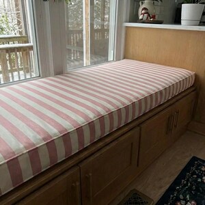 May include: A window seat cushion with a pink and white striped pattern. The cushion is rectangular and sits atop a wooden storage unit with cabinet doors. The window behind the seat offers a view of a snowy outdoor scene.