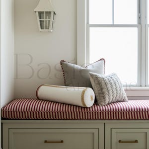May include: A window seat with a red and white striped cushion and several decorative pillows. The pillows include a gray square pillow with a red border, a white bolster pillow with gold trim, and a patterned pillow. The seat is over a cabinet with drawers.