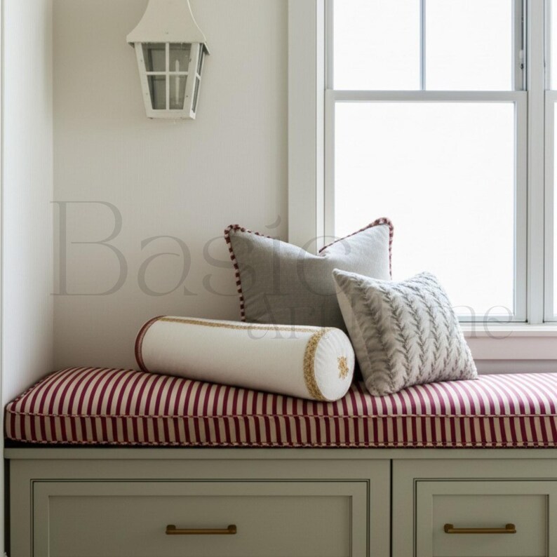 May include: A window seat with a red and white striped cushion and several decorative pillows. The pillows include a gray square pillow with a red border, a white bolster pillow with gold trim, and a patterned pillow. The seat is over a cabinet with drawers.