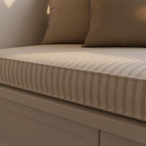 K&ouml;nnte beinhalten: Ein beiges Bankkissen mit vertikalem Streifenmuster und kontrastierendem Rand. Zwei passende beige Kissen liegen auf dem Kissen. Die Bank ist Teil einer eingebauten Sitzecke mit neutralfarbener Basis.