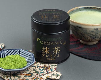 May include: A black tin of organic matcha tea with Japanese text and a bowl of matcha tea. A small blue bowl with matcha powder and a bowl of matcha tea are on a gray surface. The matcha tea is green.