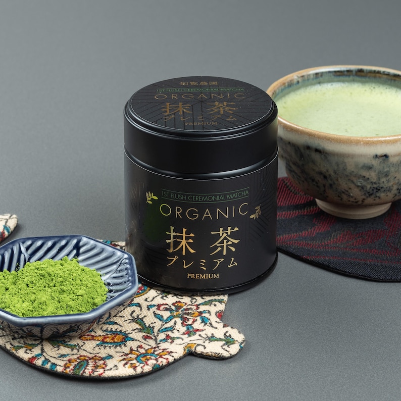 May include: A black tin of organic matcha tea with Japanese text and a bowl of matcha tea. A small blue bowl with matcha powder and a bowl of matcha tea are on a gray surface. The matcha tea is green.