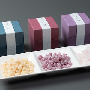 May include: Three small, square gift boxes in blue, burgundy, and purple, each with a white band and Japanese text. Below, a white rectangular dish holds three sections of star-shaped sweets in yellow, purple, and pink.