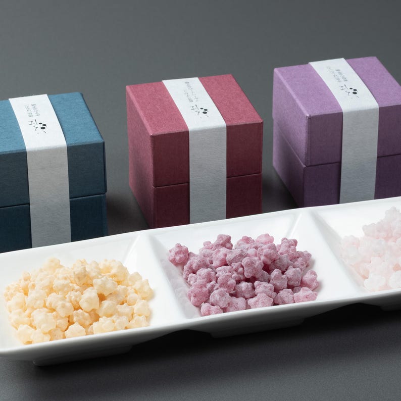 May include: Three small, square gift boxes in blue, burgundy, and purple, each with a white band and Japanese text. Below, a white rectangular dish holds three sections of star-shaped sweets in yellow, purple, and pink.