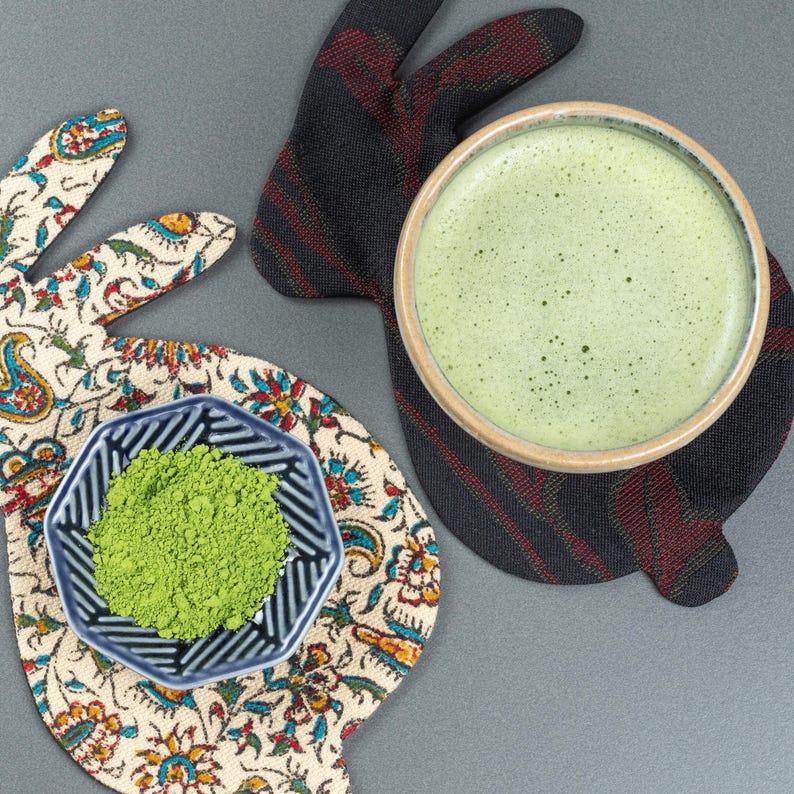 May include: A top-down view of a matcha tea set. A ceramic bowl of green matcha tea sits on a black and red plaid bunny-shaped coaster. A small blue and white patterned bowl of green matcha powder rests on a floral bunny-shaped coaster.