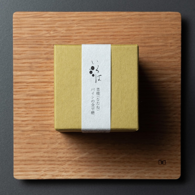 May include: A small, square, mustard-coloured box with a white label featuring Japanese text and a graphic of three black dots. The box sits on a light brown wooden cutting board, with a dark grey background.