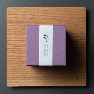 May include: A small, square, lilac box with a white vertical stripe and Japanese text, resting on a wooden cutting board. The box features Japanese text and a graphic of three black dots. The wooden board has a natural wood grain.