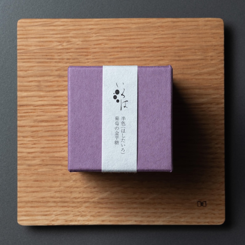 May include: A small, square, lilac box with a white vertical stripe and Japanese text, resting on a wooden cutting board. The box features Japanese text and a graphic of three black dots. The wooden board has a natural wood grain.