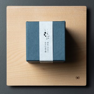 May include: A small, square, teal box with a white band, placed on a light-coloured wooden board. The box features Japanese text on the white band. The wooden board is square and has a light wood grain.