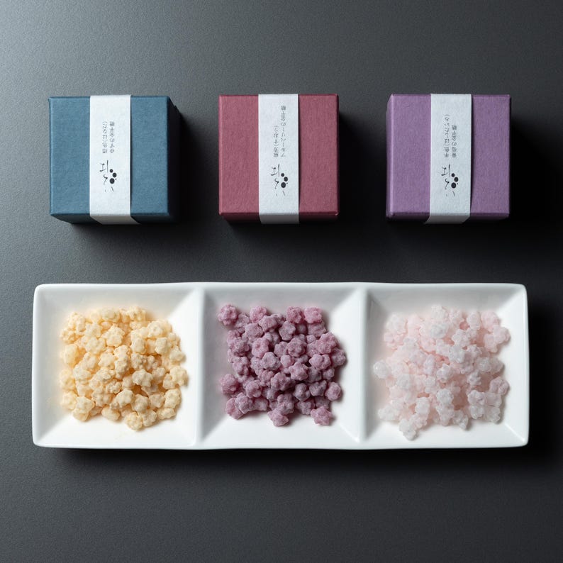 May include: Three small, square boxes in blue, burgundy, and purple, each with a white band and Japanese text. Below, a white rectangular dish holds three sections of colourful, textured sweets: yellow, purple, and pink.