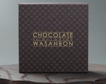 May include: A brown, checkered chocolate box with gold lettering. The text on the box reads "CHOCOLATE" and "WASANBON" in English and Japanese. The box is displayed on a dark wooden surface.