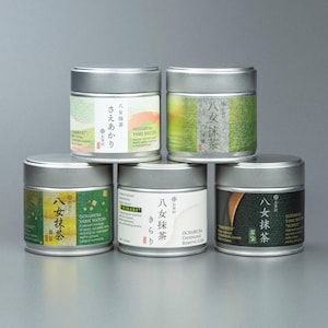 Könnte beinhalten: Fünf zylindrische Teedosen aus silbernem Metall für Ochamura Yame Matcha, die in einer Pyramide angeordnet sind. Jede Dose hat ein einzigartiges Etikett mit japanischen Schriftzeichen, englischem Text und verschiedenen Farbmustern in Grün, Gelb und Schwarz.