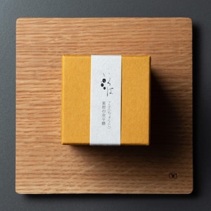 May include: A mustard-yellow box with a white label bearing Japanese text and a small illustration, placed on a wooden board. The box is square and centered, set against a dark grey background.