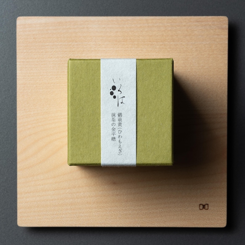 May include: An olive-green box with Japanese text on a white label, placed on a light wooden board. The board sits on a dark grey surface. The box contains a product, possibly food.