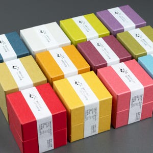 May include: A selection of brightly coloured square gift boxes. Each box is a different solid colour, such as red, yellow, green, and purple. A white band with Japanese text and a small graphic wraps around each box. The boxes are arranged on a grey surface.