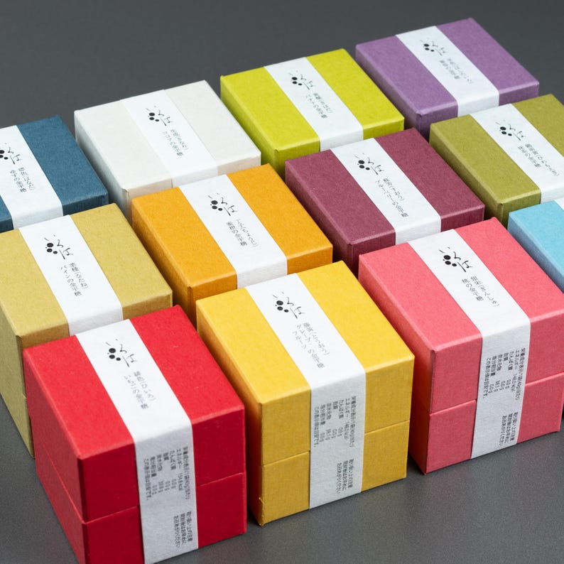May include: A selection of brightly coloured square gift boxes. Each box is a different solid colour, such as red, yellow, green, and purple. A white band with Japanese text and a small graphic wraps around each box. The boxes are arranged on a grey surface.