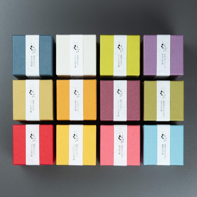 May include: Twelve small, square boxes in a variety of colours, such as blue, white, yellow, and purple. Each box features a white band with Japanese text and a small graphic. The boxes are arranged in a grid on a dark grey surface.