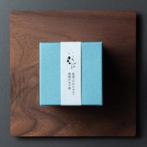 May include: A small, light blue box with a white band featuring Japanese text, placed on a dark brown wooden square. The box is centrally positioned on the wooden surface, creating a minimalist aesthetic. The wood grain is visible.