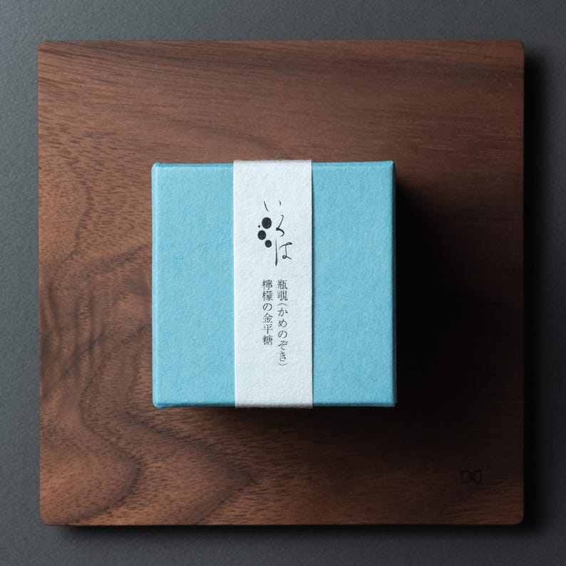 May include: A small, light blue box with a white band featuring Japanese text, placed on a dark brown wooden square. The box is centrally positioned on the wooden surface, creating a minimalist aesthetic. The wood grain is visible.