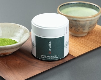 May include: A white container of Ureshino Matcha Tokunagaseicha tea sits on a wooden tray, next to a bowl of matcha powder and a bowl of matcha tea. The tea is a vibrant green color, and the bowls have a rustic, handmade appearance.