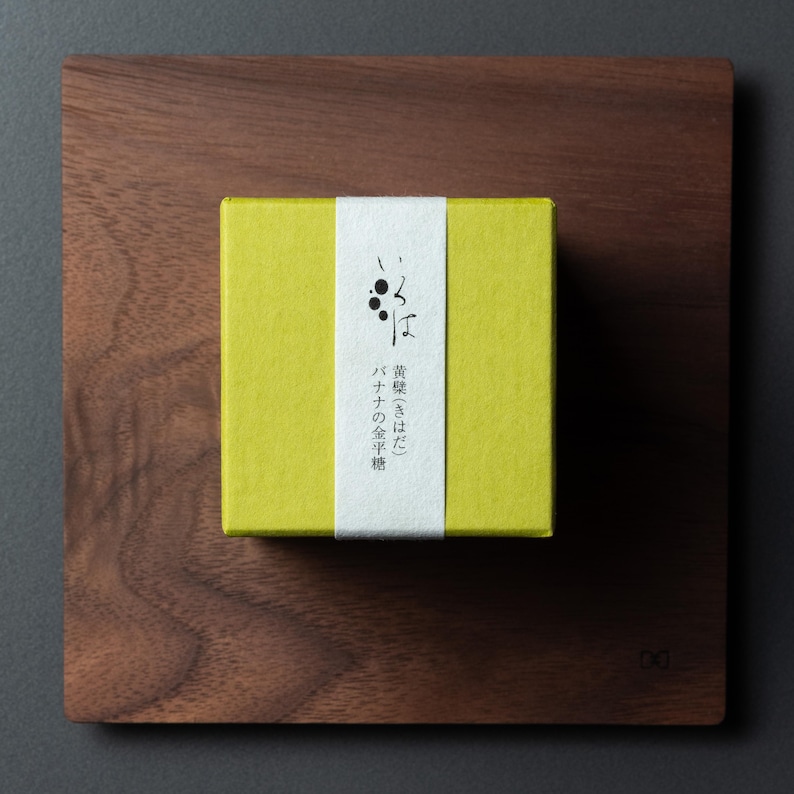 May include: A small, square, yellow box with a white band featuring Japanese text and a black graphic, on a dark wooden tray. The box likely contains a food product, such as sweets or a snack.