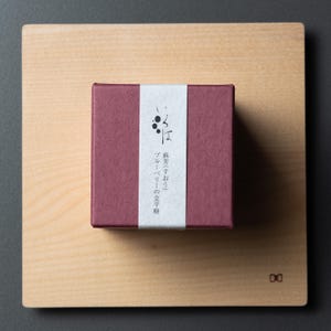 May include: A small, burgundy box with a white band featuring Japanese text and a blueberry illustration. The box sits on a light-coloured wooden square, with a minimalist design.