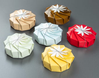 May include: Six origami-style gift boxes in various colours: tan, brown, light blue, red, yellow, and light green. Each box is octagonal with a silver ribbon and a white paper flower on top. The boxes are arranged on a grey surface.