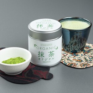Könnte beinhalten: Ein Matcha-Tee-Set mit einer weißen Schale mit grünem Matcha-Pulver, einer Metalldose mit der Aufschrift "Organic Matcha" und einer blauen Keramiktasse mit Matcha-Tee. Das Set steht auf einem gemusterten Untersetzer.