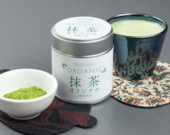 May include: An image of a matcha tea set. It includes a white ceramic bowl with green matcha powder, a metal tin labeled "Organic Matcha," and a blue ceramic cup with matcha tea. The set is on a patterned coaster.