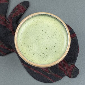 May include: A ceramic bowl filled with frothy, light green matcha tea. The bowl has a light brown rim and sits on a black and red plaid coaster. The background is a solid gray.